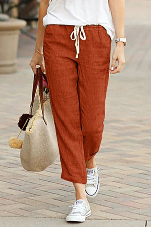 Woman wearing rust-colored relaxed fit straight pants with white drawstring and white sneakers walking outdoors carrying a beige tote bag