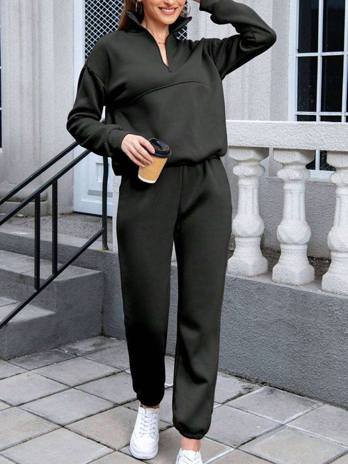 Woman wearing black cotton-blended two-piece tracksuit with high collar and zipper, holding a coffee cup outdoors.