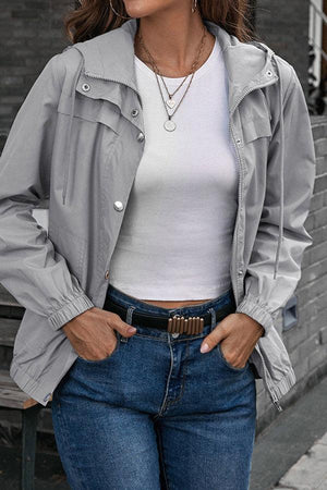 Woman wearing light grey outdoor hooded raincoat and windbreaker with white top and blue jeans
