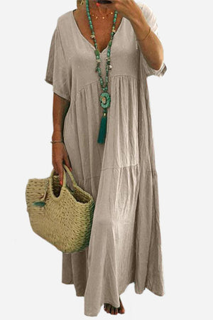 Beige V-neck two-tiered maxi swing dress with loose sleeves and flowing tiers, styled with a turquoise necklace and woven handbag