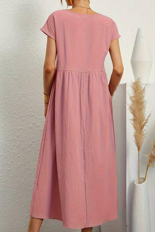 Back view of a pink capped-sleeve voluminous pleated midi dress on a woman against a white textured wall and decorative vases.