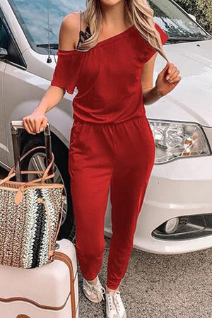 Woman wearing red Moonlit Rome Off-Shoulder jumpsuit with one shoulder exposed, standing next to a car and holding a handbag and suitcase