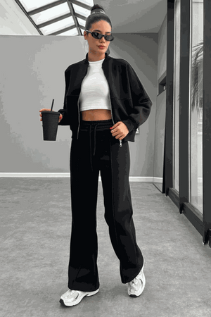 Woman wearing The Voyager Zip Set black zip-front sweatshirt and wide-leg joggers with white sneakers and sunglasses indoors.