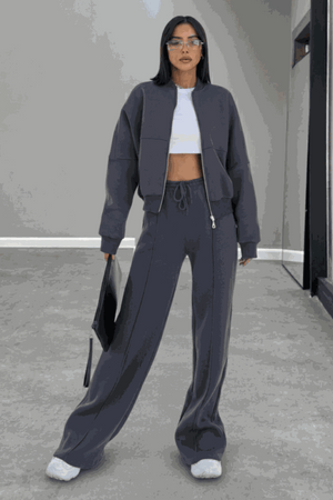 Woman wearing The Voyager Zip Set in charcoal gray, featuring a relaxed zip-front sweatshirt and wide-leg joggers with pockets, styled with white sneakers and a tote bag.