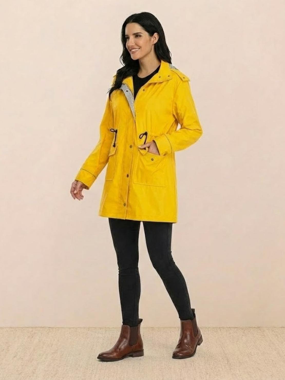 Woman wearing yellow water resistant windbreaker with hood, pockets, and adjustable waist, styled with black leggings and brown boots
