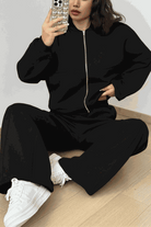 Woman wearing The Voyager Zip Set black zip-front sweatshirt and wide-leg joggers with white sneakers sitting on floor taking a mirror selfie