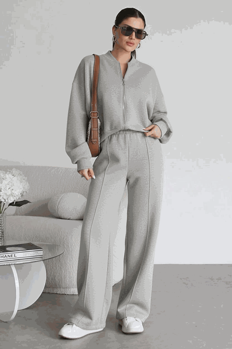 Woman wearing The Voyager Zip Set grey zip-front sweatshirt and wide-leg joggers with pockets, styled with sunglasses, white sneakers, and a brown tote bag in a modern indoor setting.
