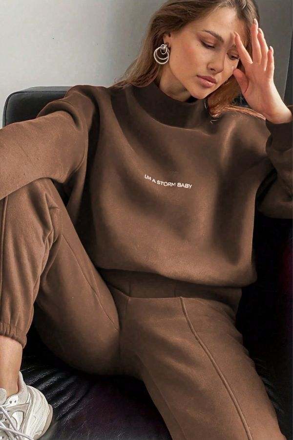 Woman wearing a brown Storm Sweater Mock Neck athleisure set with relaxed fit and matching joggers sitting on a sofa.