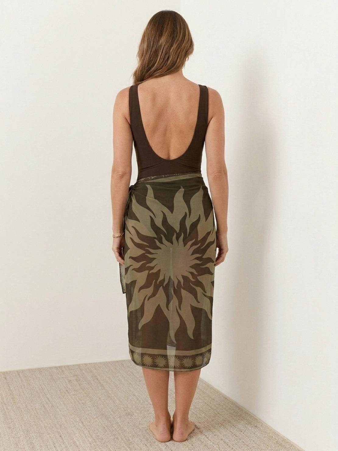 One-piece deep-V swimsuit with ruched bust and waist, wide straps, gold shell detail, low back, high-cut leg, and sheer celestial print sarong wrap.