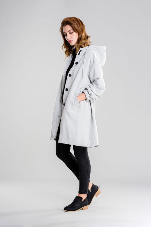 Woman wearing a light gray water-resistant oversized hooded windbreaker rain jacket with black buttons and black leggings.