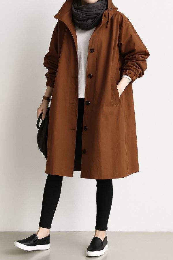 Woman wearing brown water resistant oversized windbreaker rain jacket with hood and button closure, styled with black pants and slip-on shoes.
