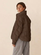 Woman wearing brown lightweight puffer travel jacket with high collar and gray pants, side view.