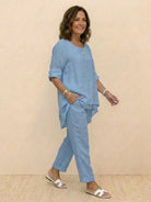Woman wearing a light blue asymmetrical two-piece trouser set with rolled sleeves and tapered pants, styled casually with white sandals.
