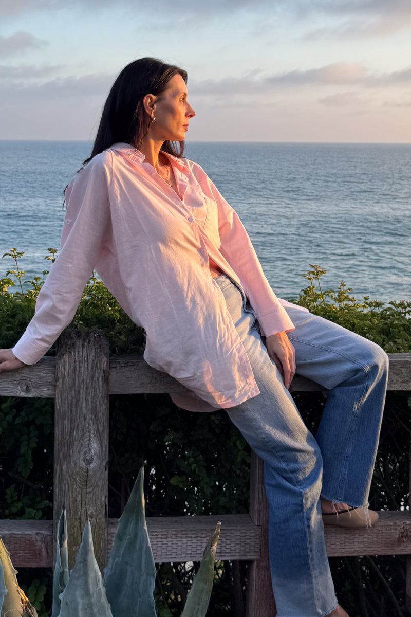 Woman wearing oversized linen shirt in soft pink, sitting by ocean at sunset