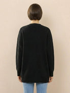 Back view of woman wearing black relaxed fit oversized button down knitwear cardigan with long sleeves and round neckline paired with light blue jeans against beige background