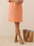 Woman wearing peach knee-length cotton dress with pockets and rolled 3/4 sleeves, standing next to woven tote bag and tan sandals